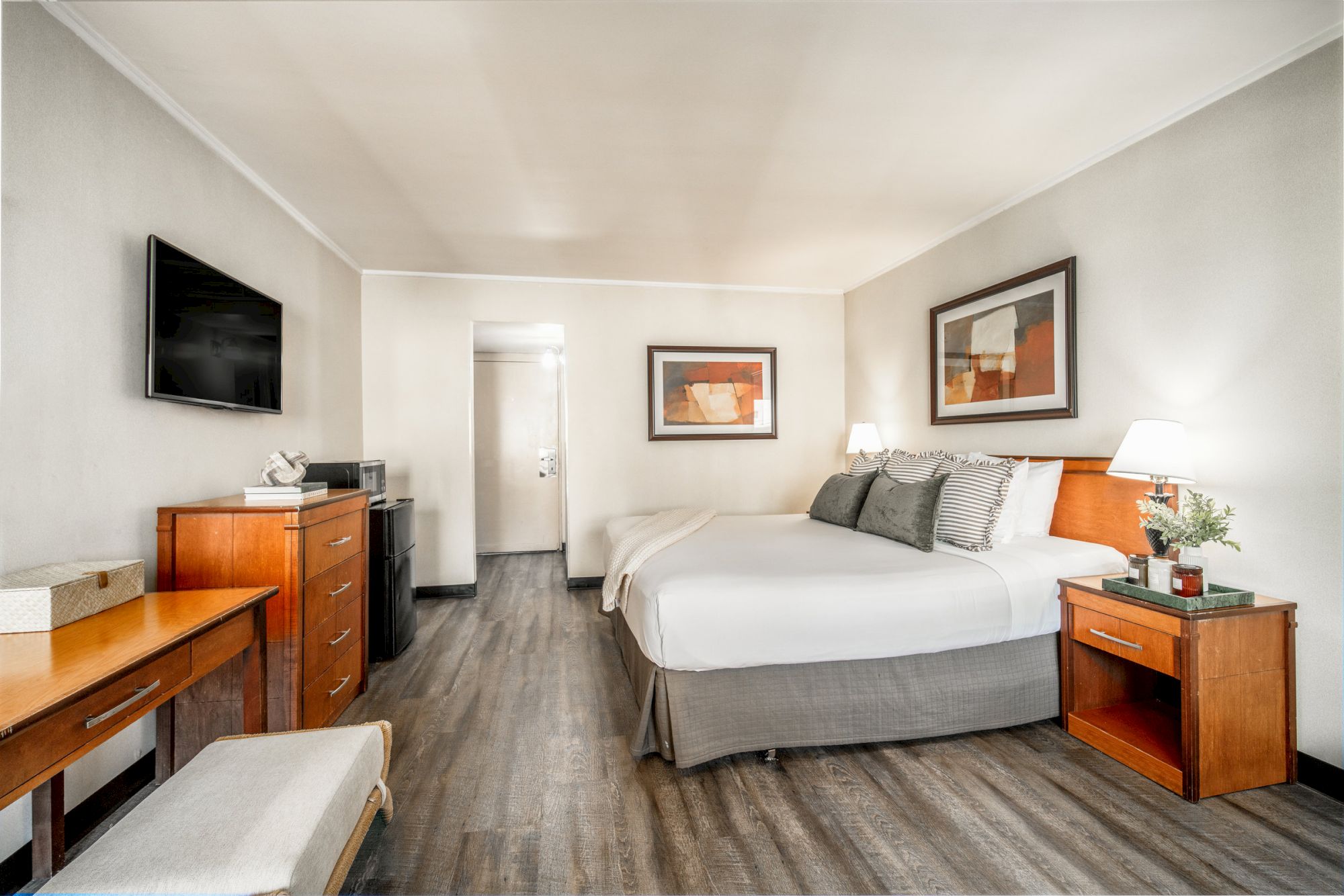 A tidy hotel bedroom with a king-size bed, flat-screen TV, dresser, nightstands, lamps, and framed abstract art.