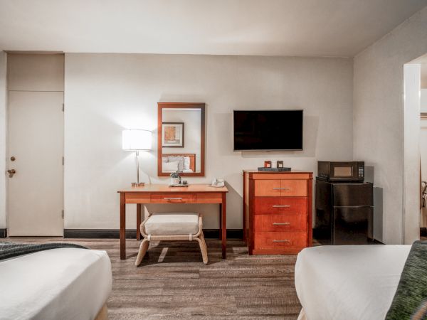 A bright hotel-style room with a desk and lamp, a TV on the wall, an orange dresser, and a kitchenette with microwave and fridge.