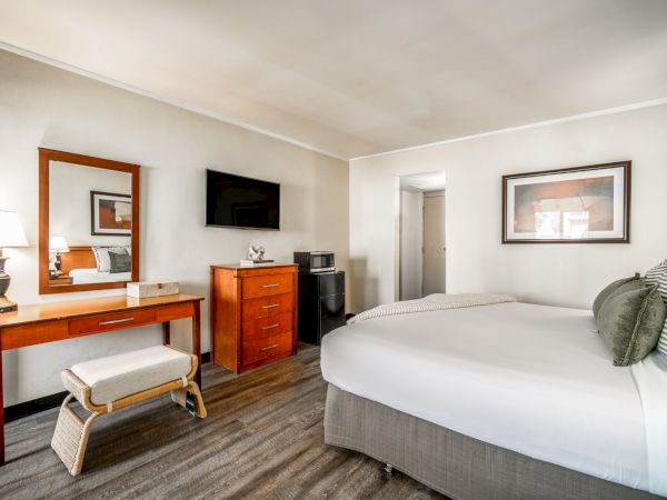 A bright hotel room with a bed, TV, dresser, framed art, and a mirror.