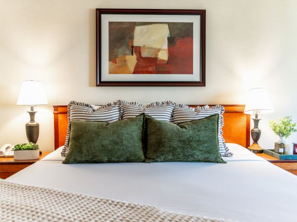 A cozy bedroom scene with a bed, striped pillows, two lamps, and a framed abstract artwork on the wall.