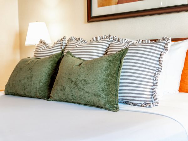 A neatly made bed with green cushions, striped pillows, a floor lamp, and framed wall art in a warm room.