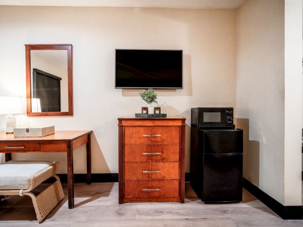 A hotel room corner with a wooden dresser, wall TV, microwave, black refrigerator, lamp, mirror, and small table.