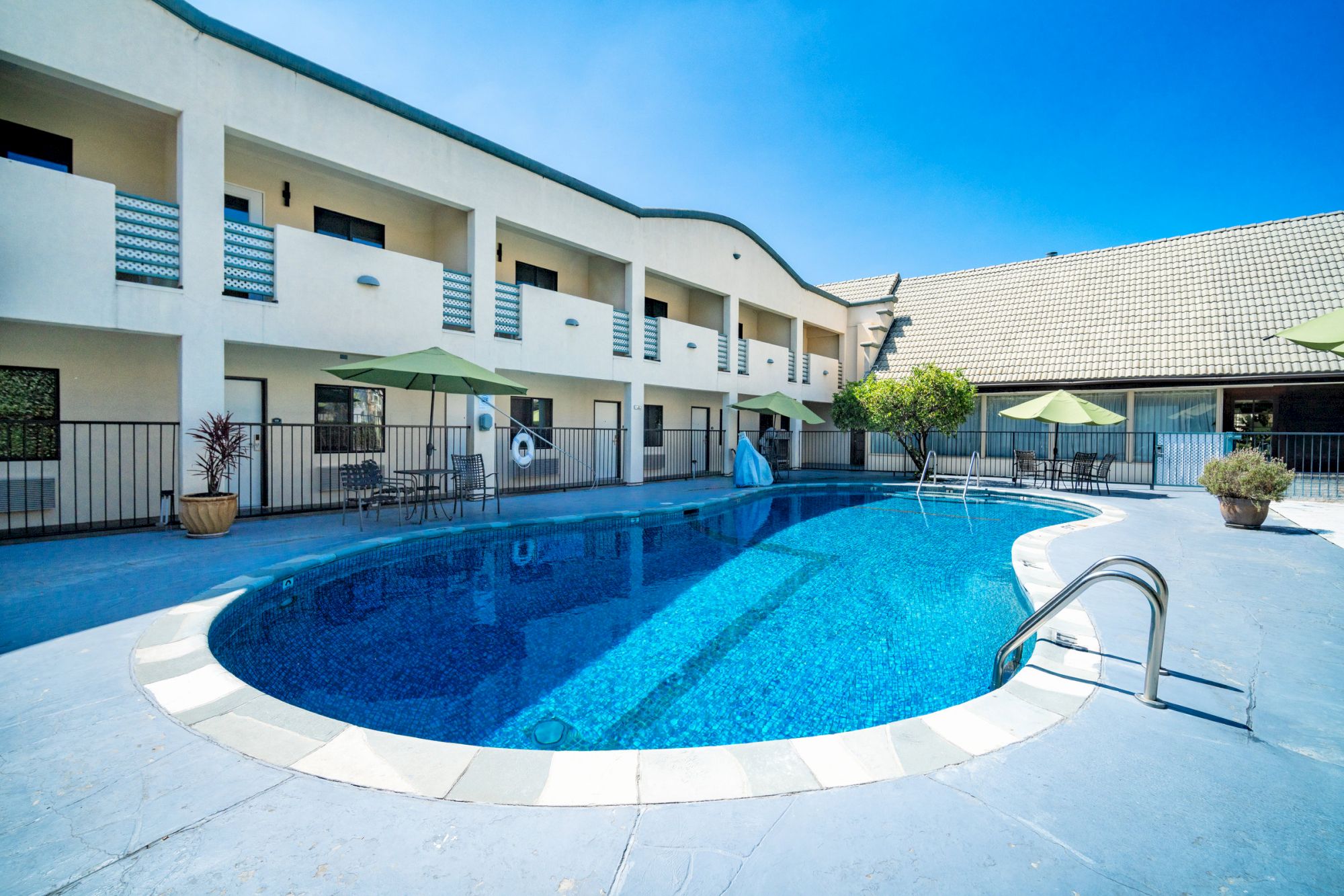 A modern courtyard with a tiled swimming pool, lounge chairs, green umbrellas, and white apartment buildings around it.